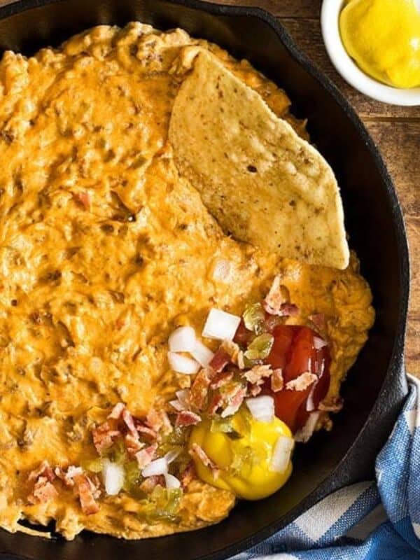 A skillet filled with creamy cheeseburger dip, topped with chopped onions, tomatoes, bacon, and mustard, surrounded by tortilla chips, ketchup, mustard, relish, and a blue-striped towel on a wooden table.