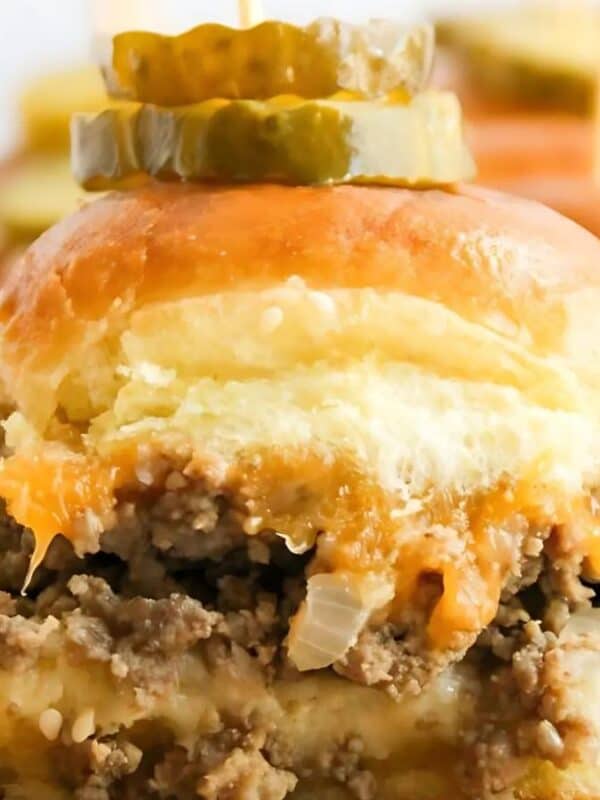 A close-up of a cheesy ground beef slider on a soft bun, topped with pickle slices. Other similar sliders are blurred in the background.