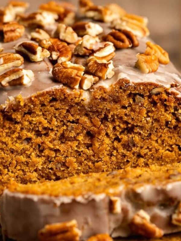 A loaf of glazed pumpkin bread topped with chopped pecans, with one slice cut and laying in front. The bread looks moist and is placed on parchment paper.