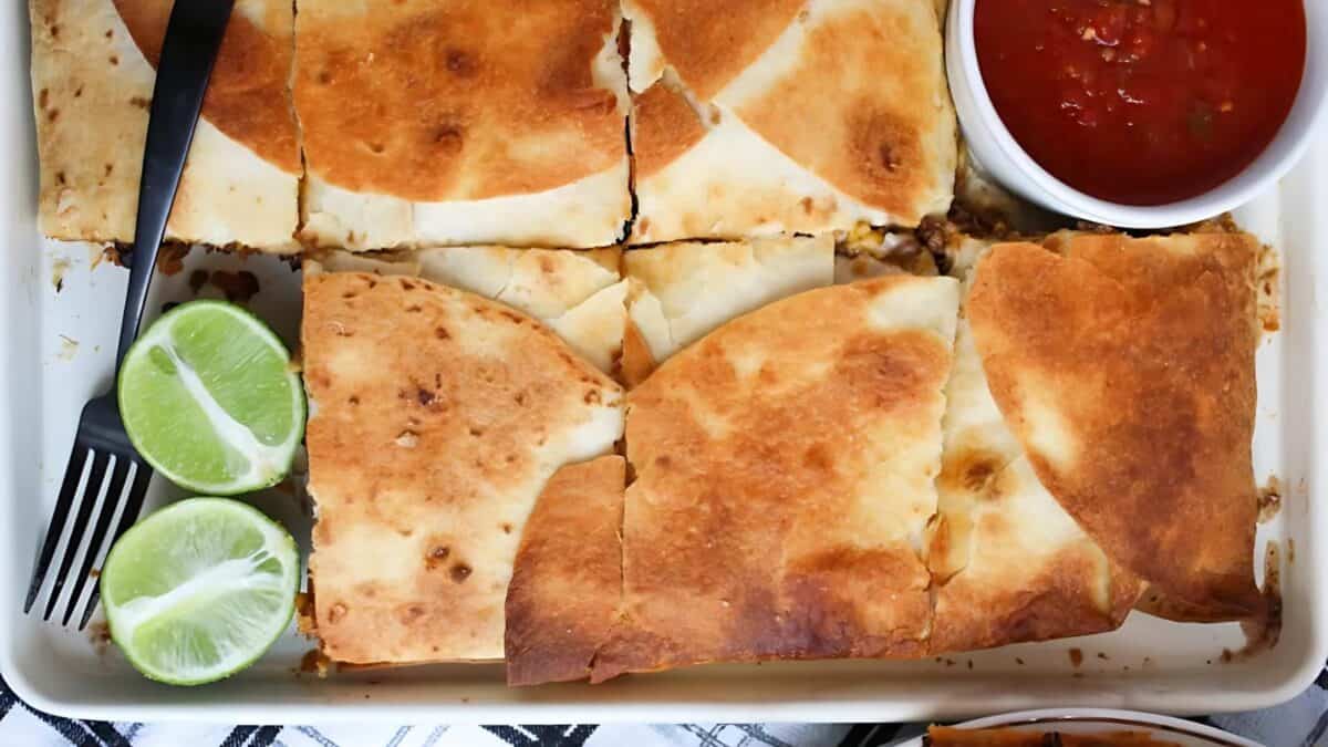 A baking dish with golden, baked quesadillas cut into squares, served with lime wedges and a small bowl of red salsa. A black spatula is beside the quesadillas.
