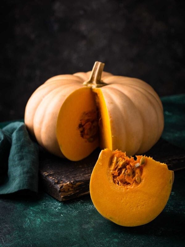 A whole pumpkin with light orange skin sits on a dark surface, with a large wedge cut out to reveal its bright orange flesh and seeds. A green cloth is nearby, and the background is dark and textured.