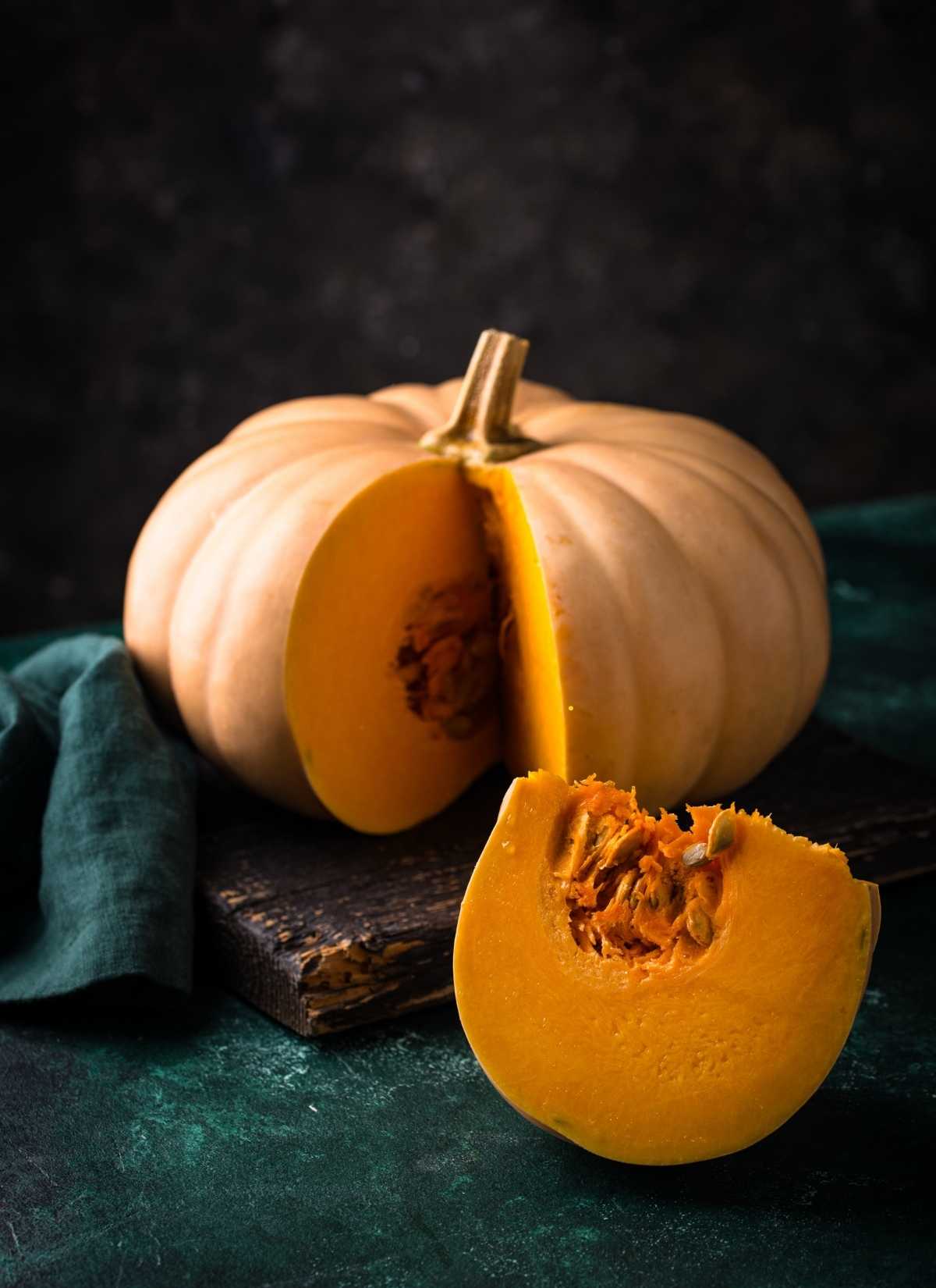 A whole pumpkin with light orange skin sits on a dark surface, with a large wedge cut out to reveal its bright orange flesh and seeds. A green cloth is nearby, and the background is dark and textured.