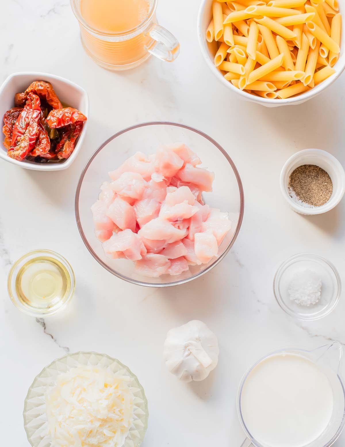 Overhead view of neatly arranged ingredients: cubed raw chicken, penne pasta, grated cheese, sun-dried tomatoes, garlic, cream, broth, oil, salt, and pepper on a white marble surface.