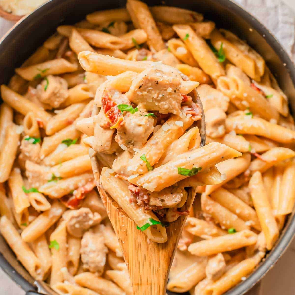 A close-up of a wooden spoon lifting creamy penne pasta with chicken, sun-dried tomatoes, and fresh herbs from a large skillet filled with the same pasta dish.