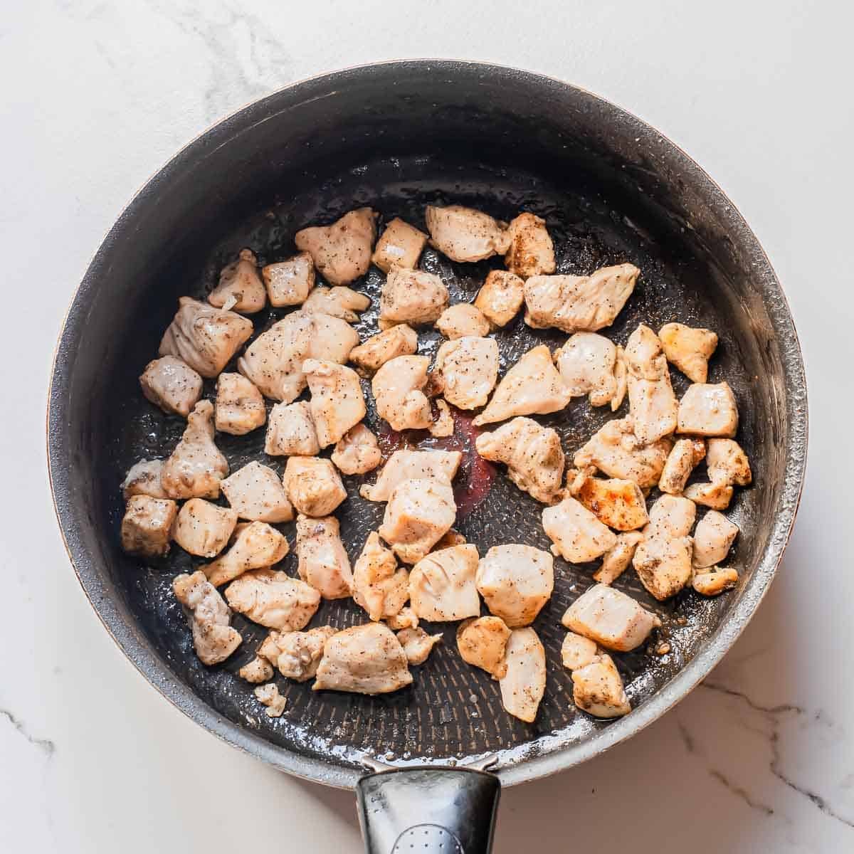 Cubed pieces of seasoned chicken are cooking in a black frying pan on a white marble surface. The chicken is lightly browned and spread evenly across the pan.