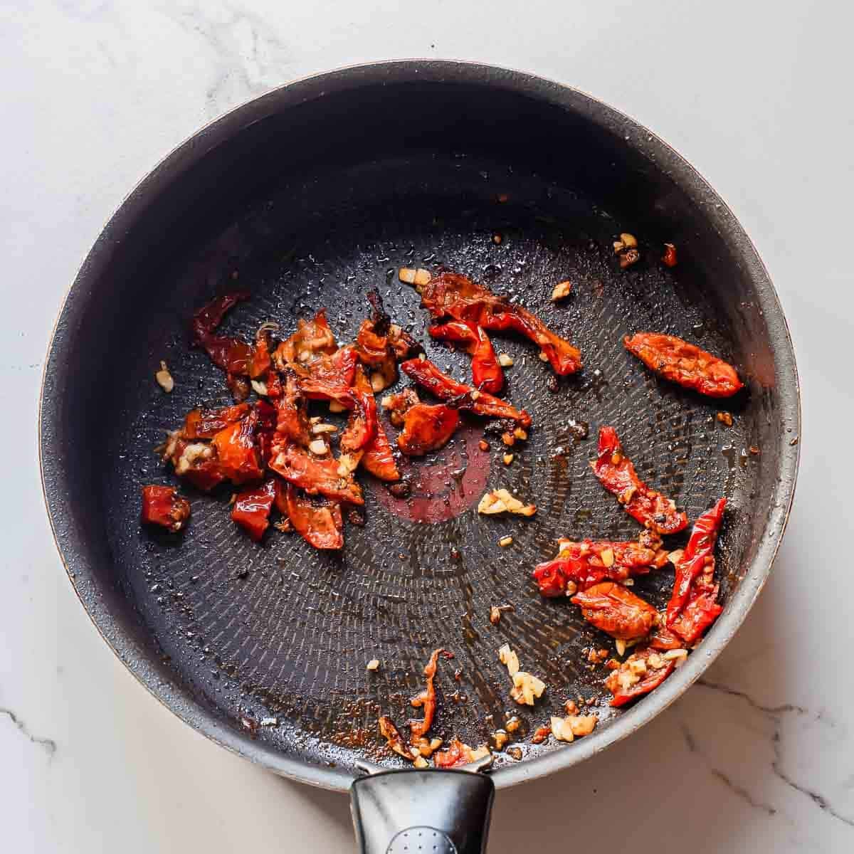 A black frying pan on a white surface contains sautéed chopped sun-dried tomatoes and minced garlic, lightly browned and scattered across the pan.