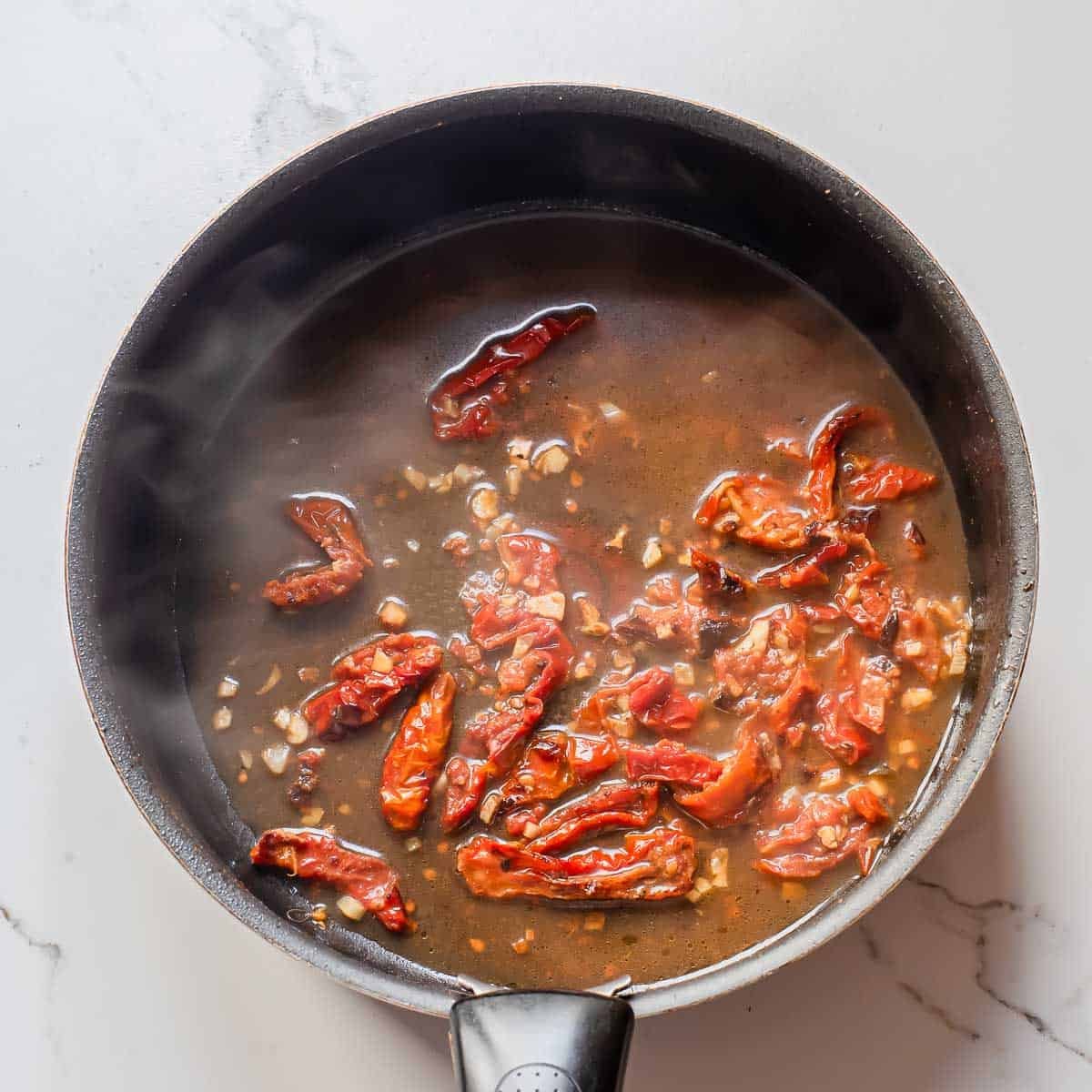 A black frying pan on a light countertop contains a simmering mixture of sun-dried tomatoes, chopped garlic, and liquid. Steam rises from the pan, indicating the ingredients are being cooked.
