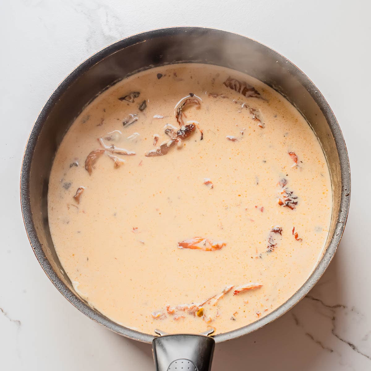 A black saucepan filled with a creamy, orange-colored soup containing chunks of shredded meat and mushrooms, set on a white marble surface. Steam is rising from the hot soup.