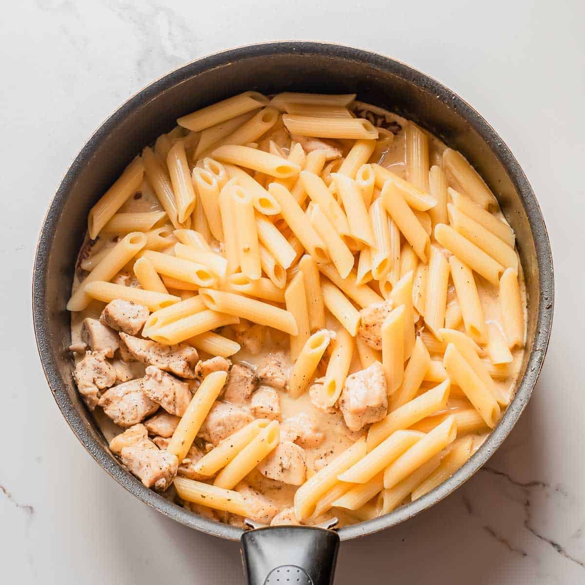 A black frying pan on a white surface contains cooked penne pasta and pieces of seasoned chicken in a creamy sauce.