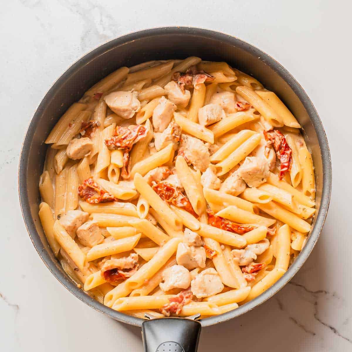 A skillet filled with cooked penne pasta, diced chicken, and sun-dried tomatoes in a creamy sauce, viewed from above on a light surface.