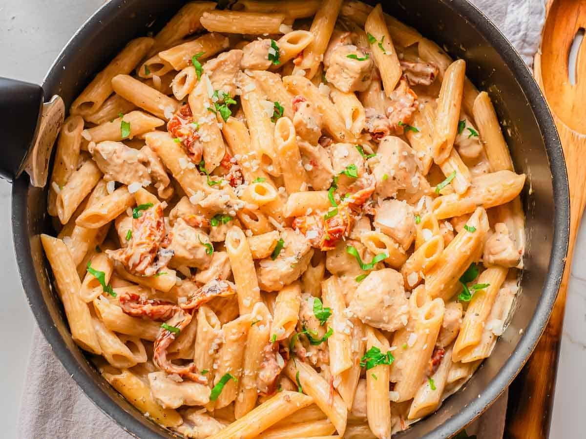 A skillet filled with marry me creamy chicken penne pasta and garnished with chopped parsley and crumbled cheese.