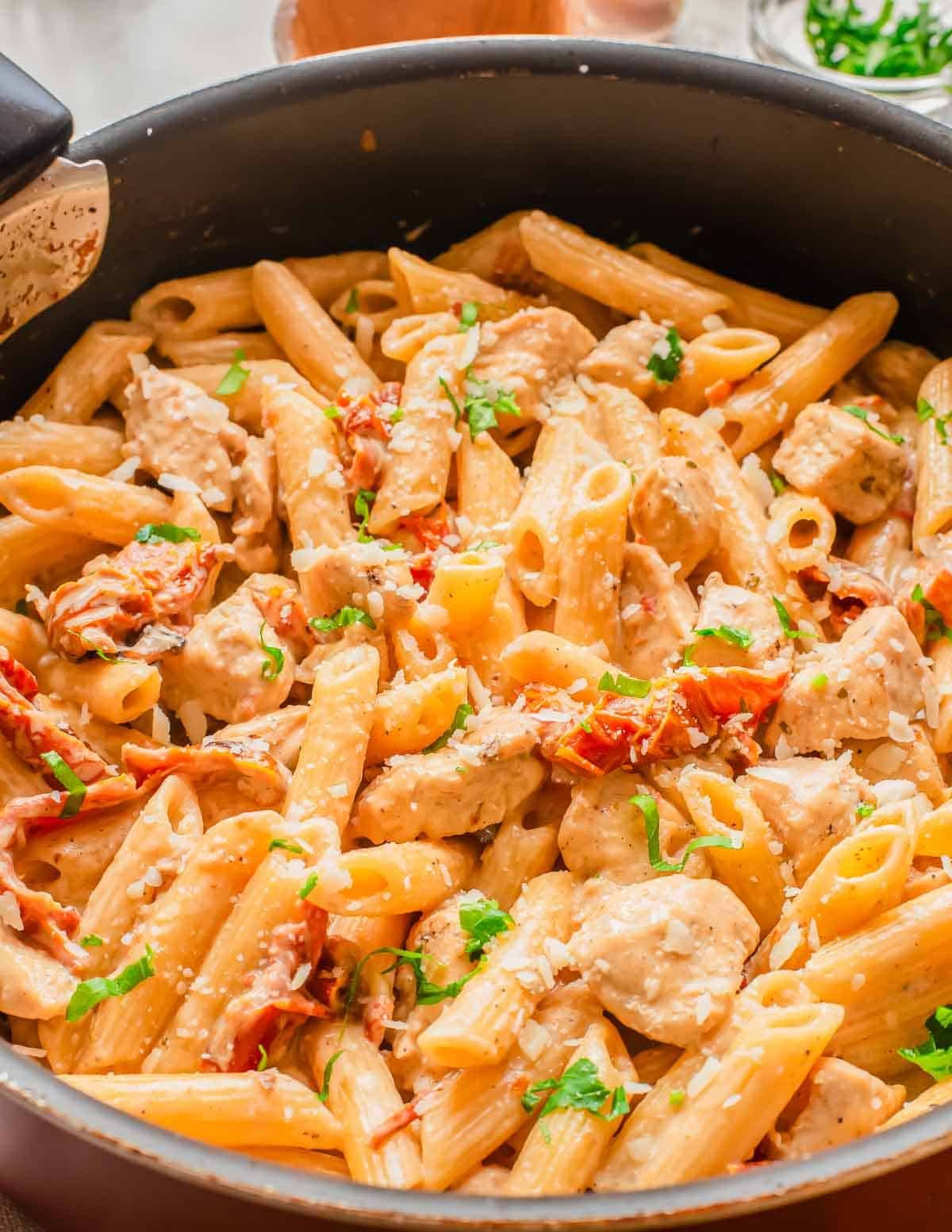 A skillet filled with creamy penne pasta, chunks of chicken, sun-dried tomatoes, and garnished with chopped parsley and grated cheese.
