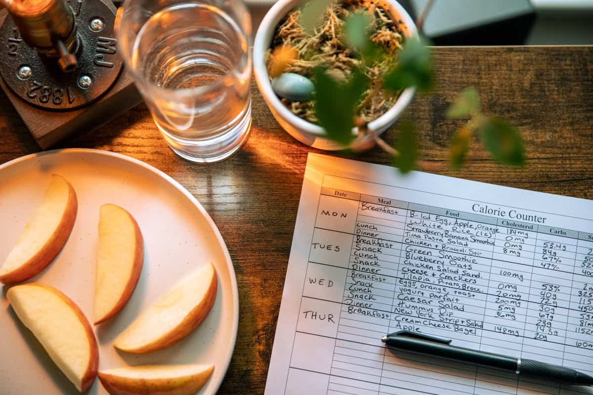 A plate with apple slices, a glass of water, a potted plant, and a calorie counter chart with a pen on a wooden table, tracking meals and calories for the week.