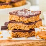 Three stacked peanut butter oat and chocolate bars with a thick chocolate topping and flaky salt, resting on a wooden board. Crumbs and a dollop of peanut butter are visible nearby. More bars are blurred in the background.
