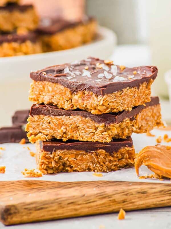 Three stacked peanut butter oat and chocolate bars with a thick chocolate topping and flaky salt, resting on a wooden board. Crumbs and a dollop of peanut butter are visible nearby. More bars are blurred in the background.