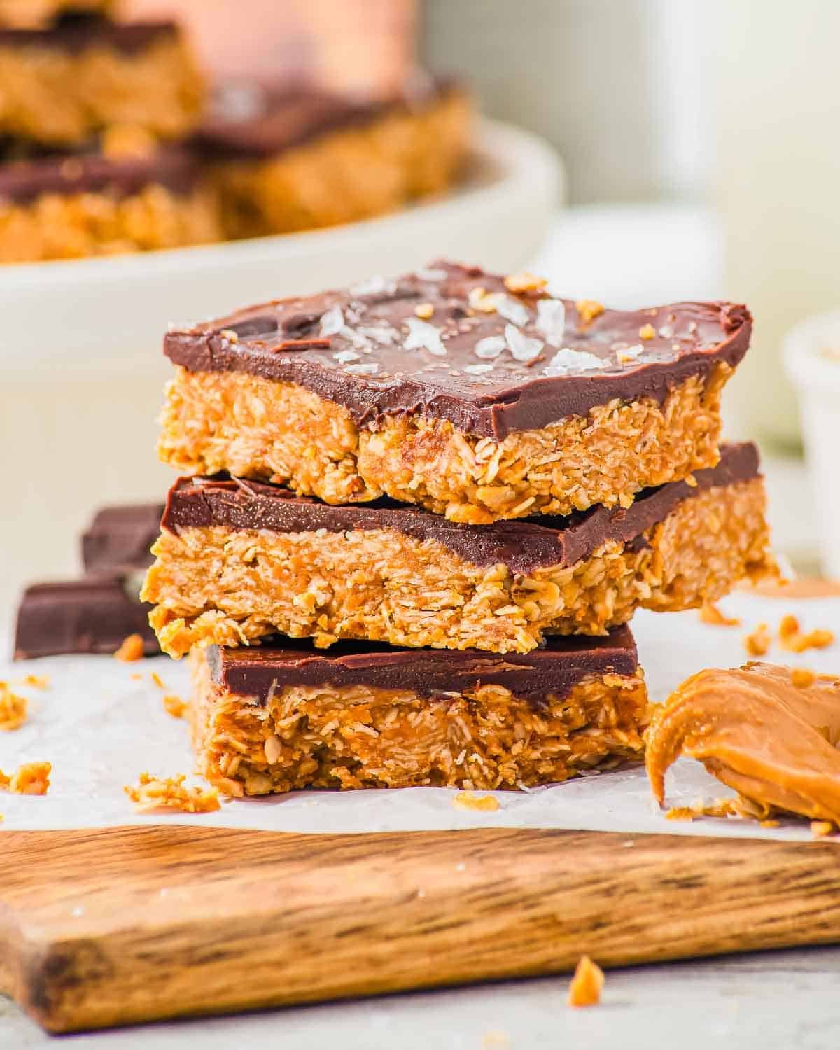 Three stacked peanut butter oat and chocolate bars with a thick chocolate topping and flaky salt, resting on a wooden board. Crumbs and a dollop of peanut butter are visible nearby. More bars are blurred in the background.