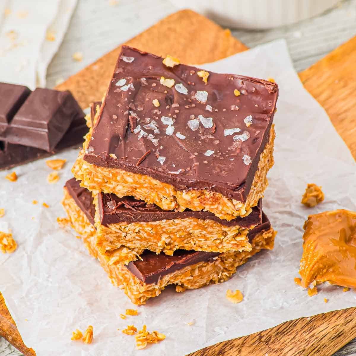 Three oatmeal peanut butter and chocolate bars stacked on parchment paper, topped with flaky sea salt. A piece of chocolate and some caramel spread are nearby on a wooden board.