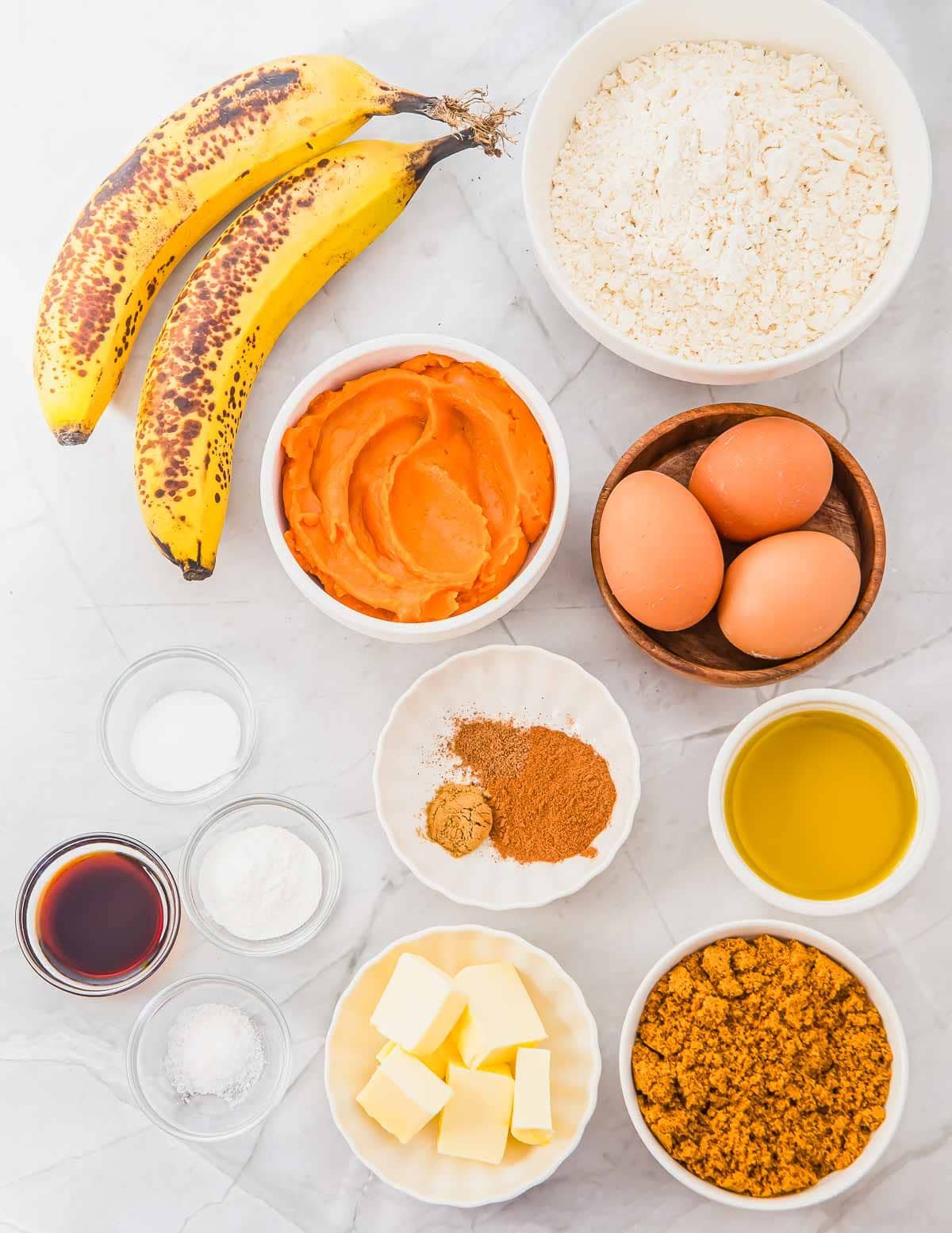 A top-down view of baking ingredients on a white surface, including ripe bananas, flour, pumpkin puree, eggs, oil, brown sugar, butter, vanilla extract, spices, baking powder, and salt in bowls.