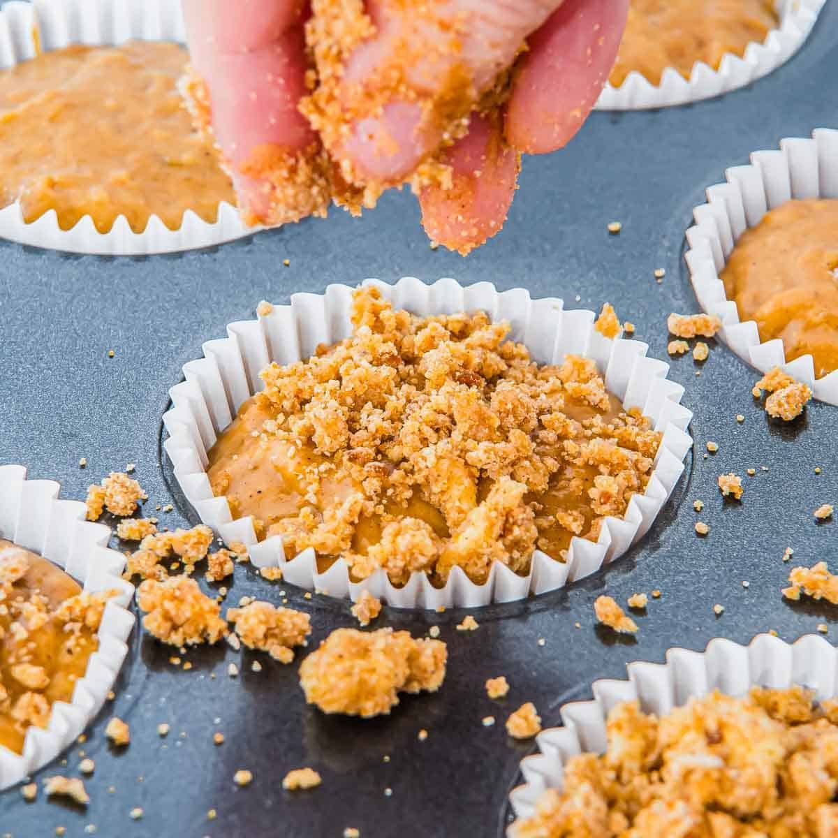 A hand sprinkles crumb topping onto pumpkin banana muffin batter in a lined muffin tin, with other unbaked muffins nearby.