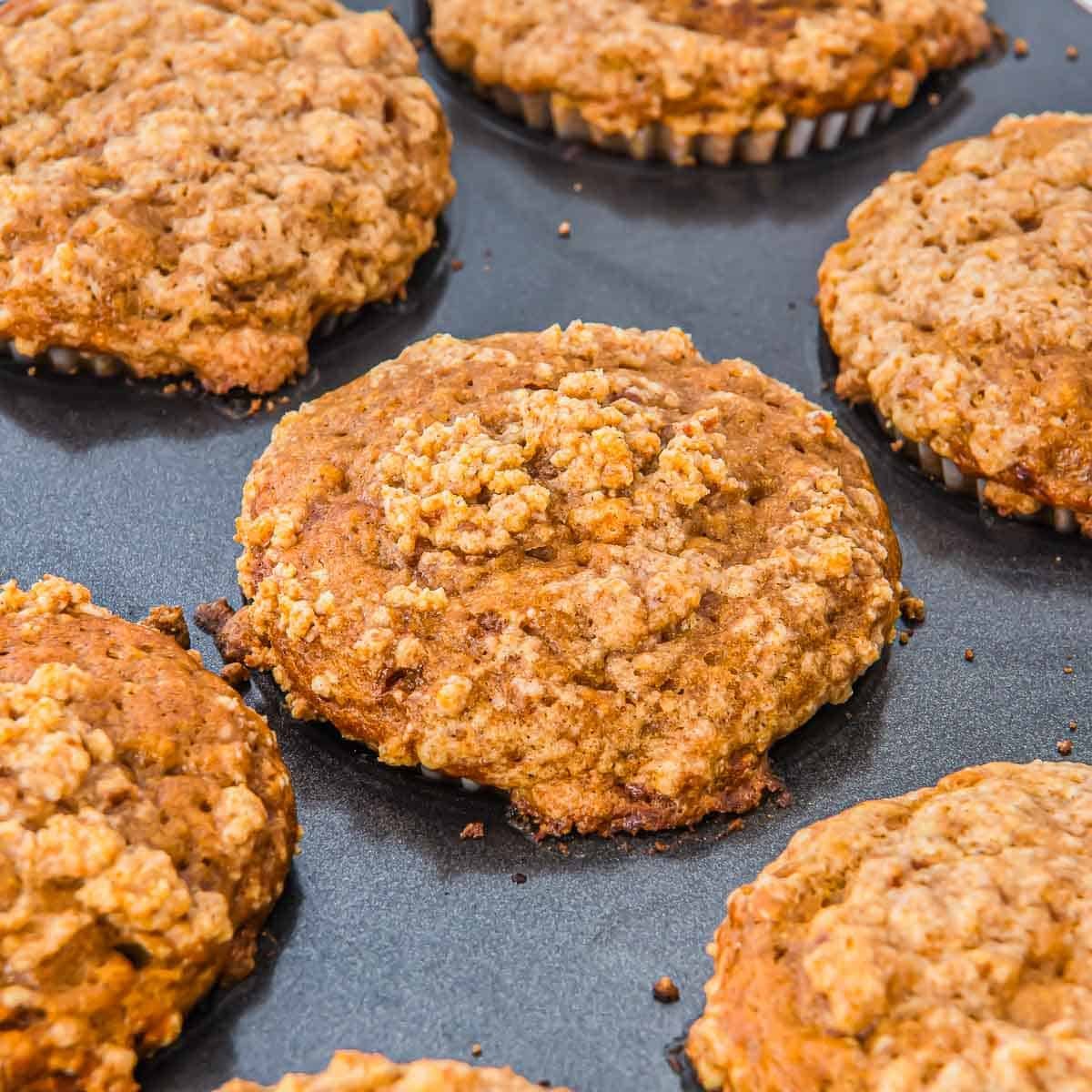 Close-up of freshly baked pumpkin banan muffins with a crumbly streusel topping, arranged on a dark baking tray. The muffins appear golden brown and textured, suggesting a homemade, rustic style.