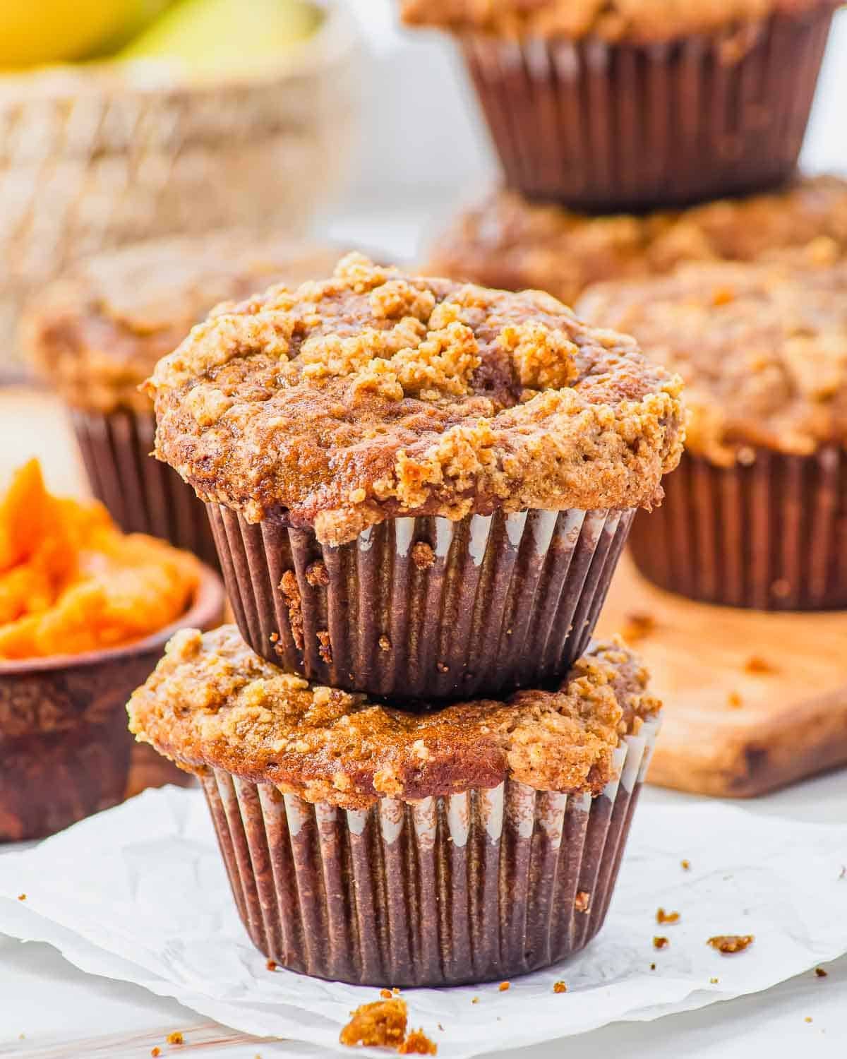Two pumpkin banana muffins with streusel topping are stacked on parchment paper, with more muffins and a bowl of pumpkin puree in the background, creating a cozy and inviting scene.
