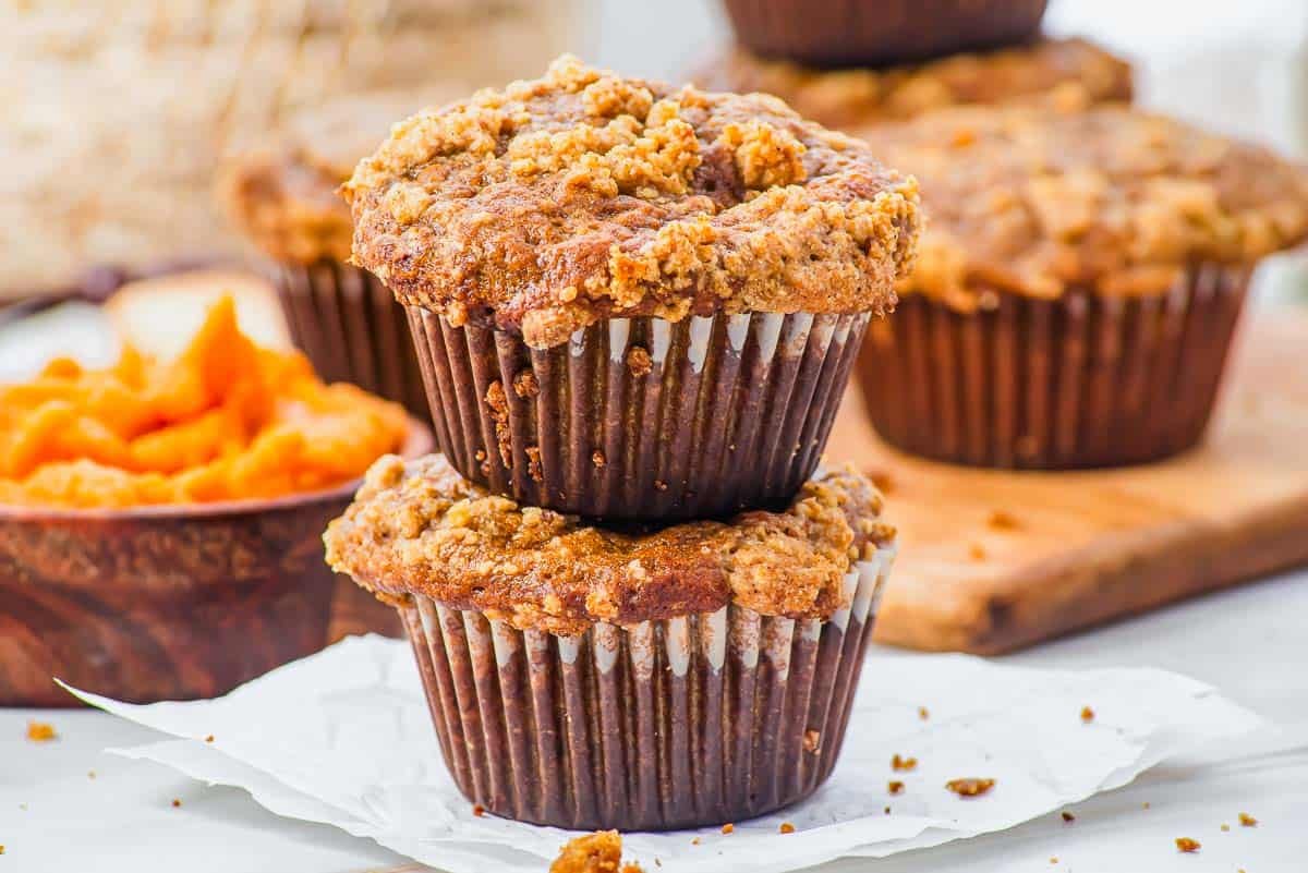 Two crumb-topped pumpkin banana muffins stacked on each other, resting on crumpled parchment paper, with more muffins and a bowl of pumpkin filling blurred in the background.