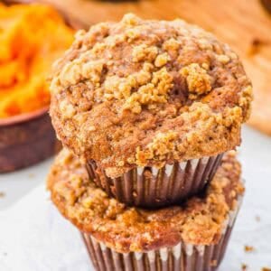 Two pumpkin banana muffins with a crumbly streusel topping are stacked on top of each other on a piece of parchment paper, with a bowl of pumpkin and more muffins in the background.