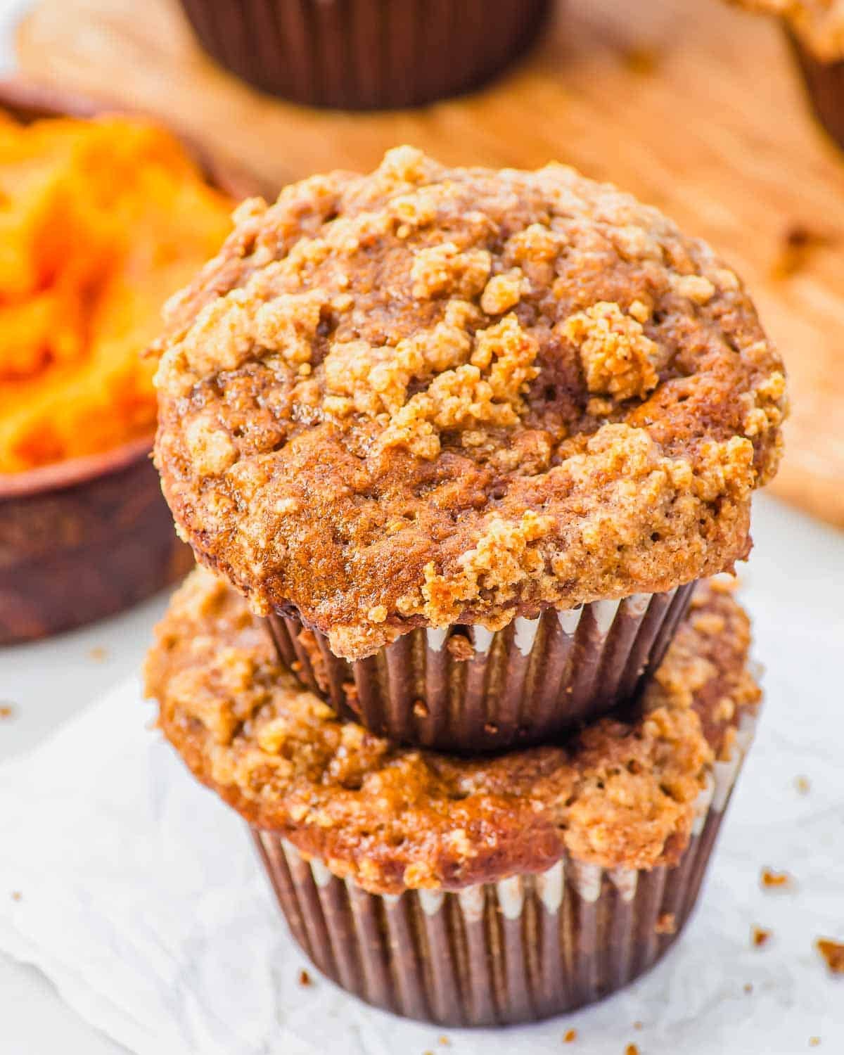 Two pumpkin banana muffins with a crumbly streusel topping are stacked on top of each other on a piece of parchment paper, with a bowl of pumpkin and more muffins in the background.