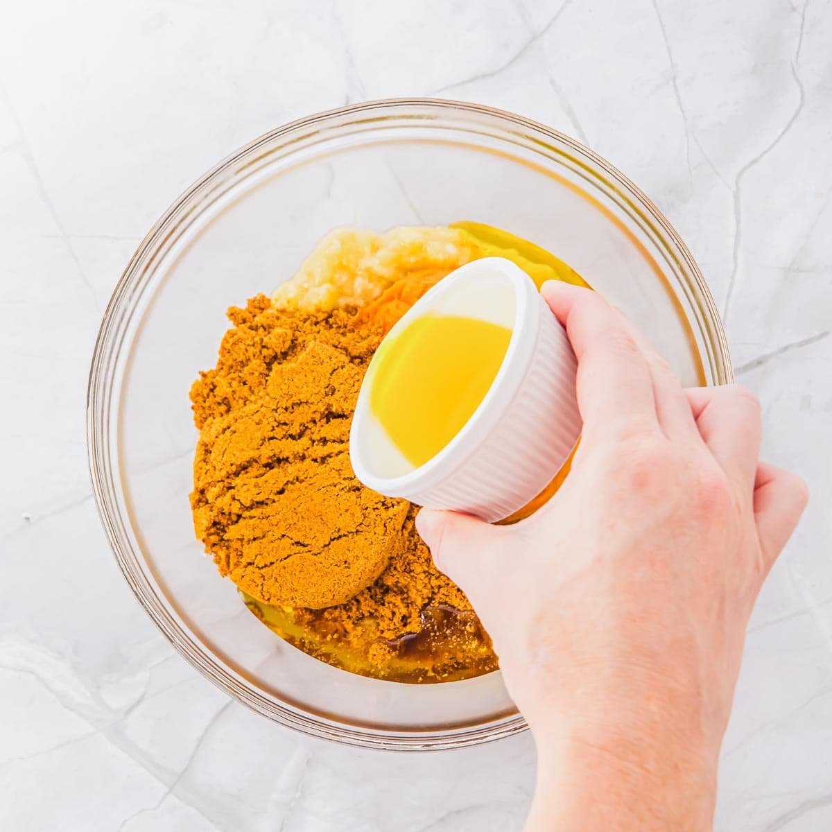 A hand pours oil from a small white cup into a glass bowl containing mashed bananas, brown sugar, and other baking ingredients on a white marble surface.