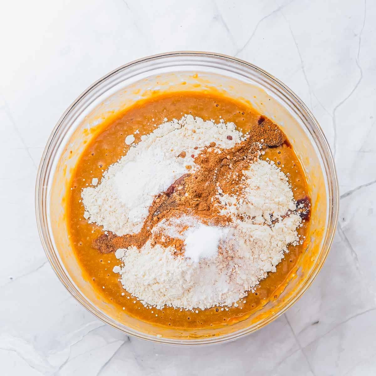 A glass bowl on a marble surface filled with pumpkin batter, topped with piles of flour, baking soda, baking powder, spices, and salt, ready to be mixed.