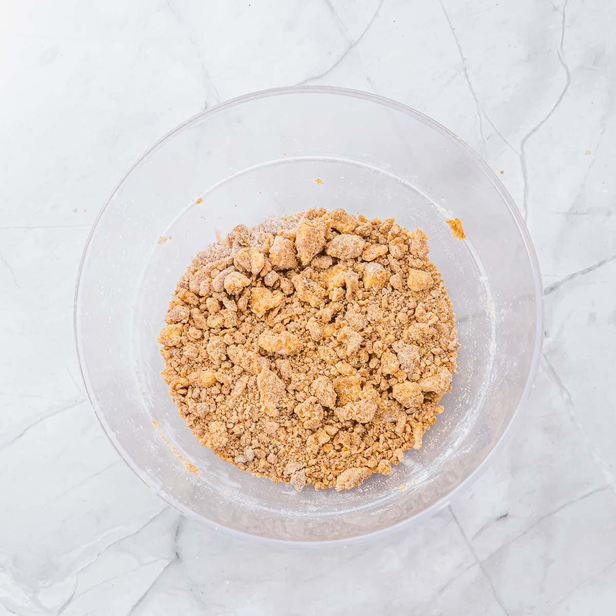 A clear glass bowl filled with crumbly brown streusel mixture sits on a white marble surface.