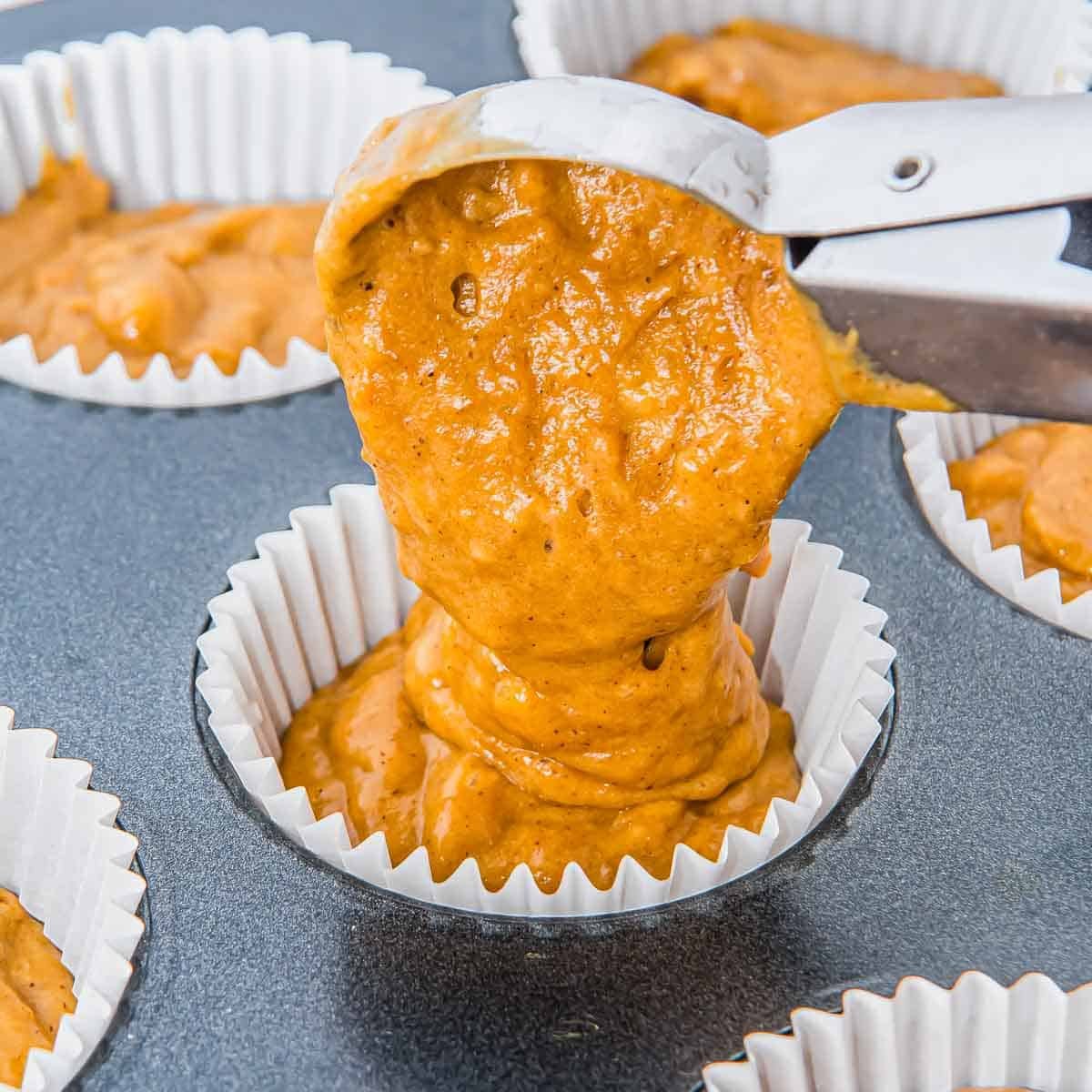 A metal ice cream scoop fills a white paper muffin liner with pumpkin banana muffin batter. Other liners in the tray are already partially filled with the same batter.