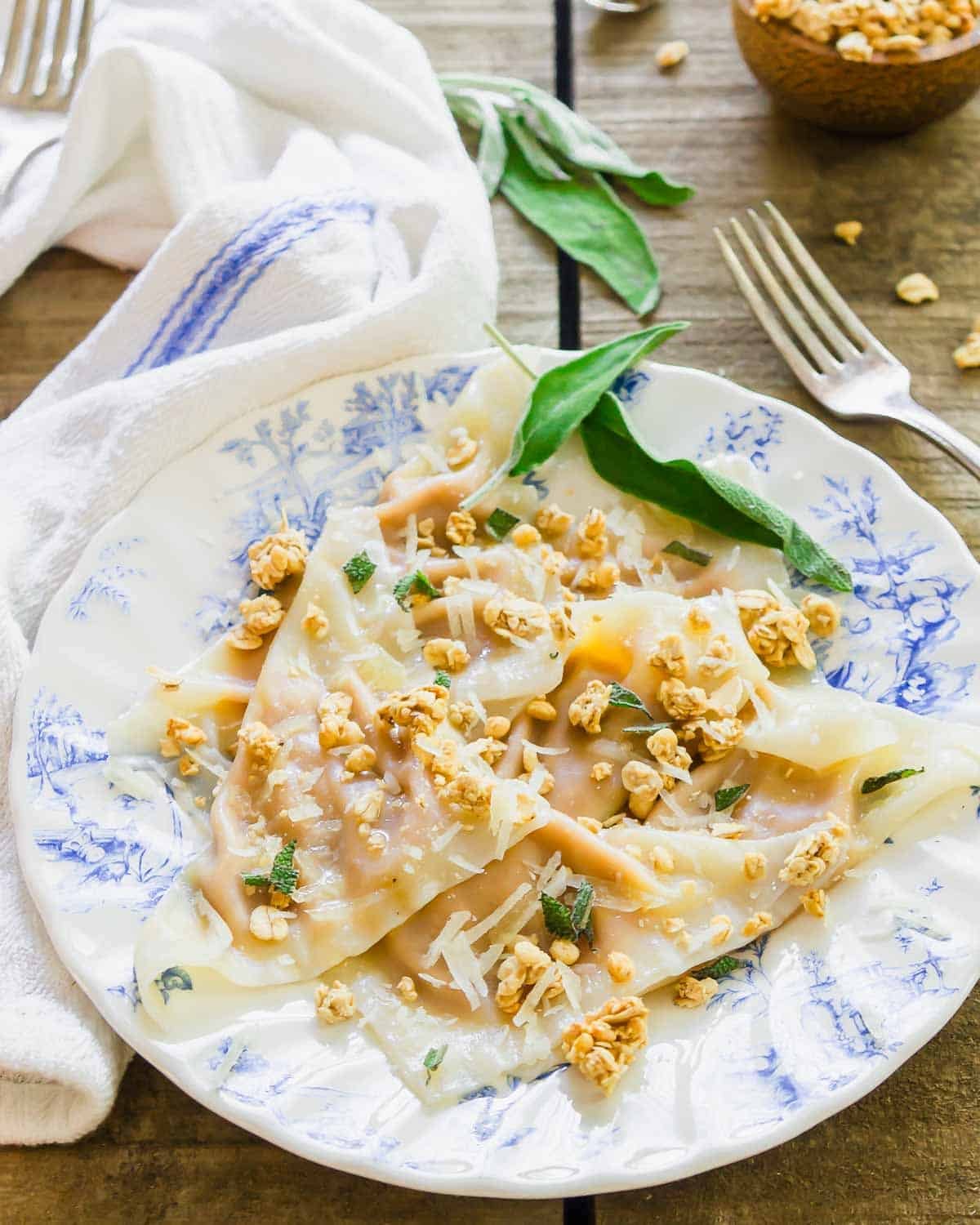 A floral-patterned plate holds ravioli topped with grated cheese, fresh sage, and crunchy granola. A fork, white towel, and scattered granola pieces are nearby on a rustic wooden table.