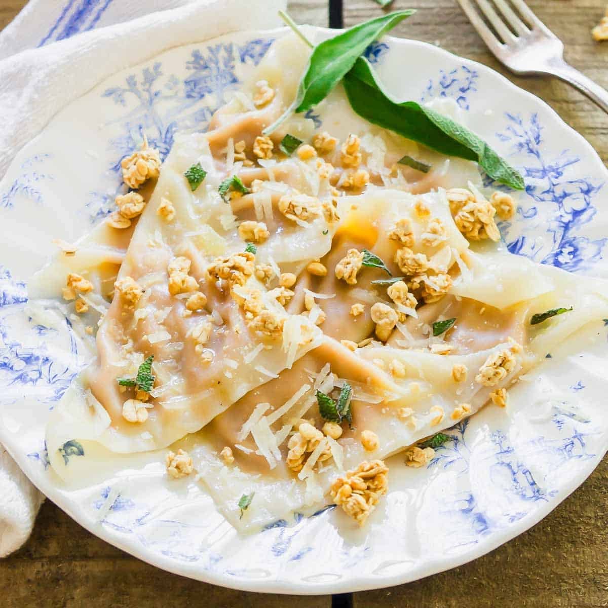 A plate with three folded ravioli topped with grated cheese, crispy sage, and crunchy bits, served on a blue and white patterned dish with a fork and cloth napkin nearby.