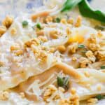 Close-up of ravioli topped with grated cheese, fresh herbs, and crunchy granola on a decorative plate. A green leafy garnish is placed beside the pasta.