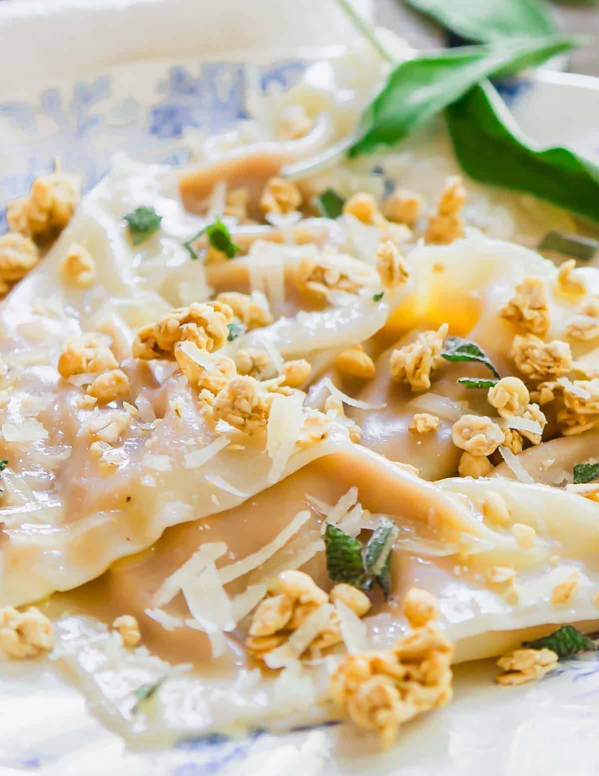 Close-up of ravioli topped with grated cheese, fresh herbs, and crunchy granola on a decorative plate. A green leafy garnish is placed beside the pasta.