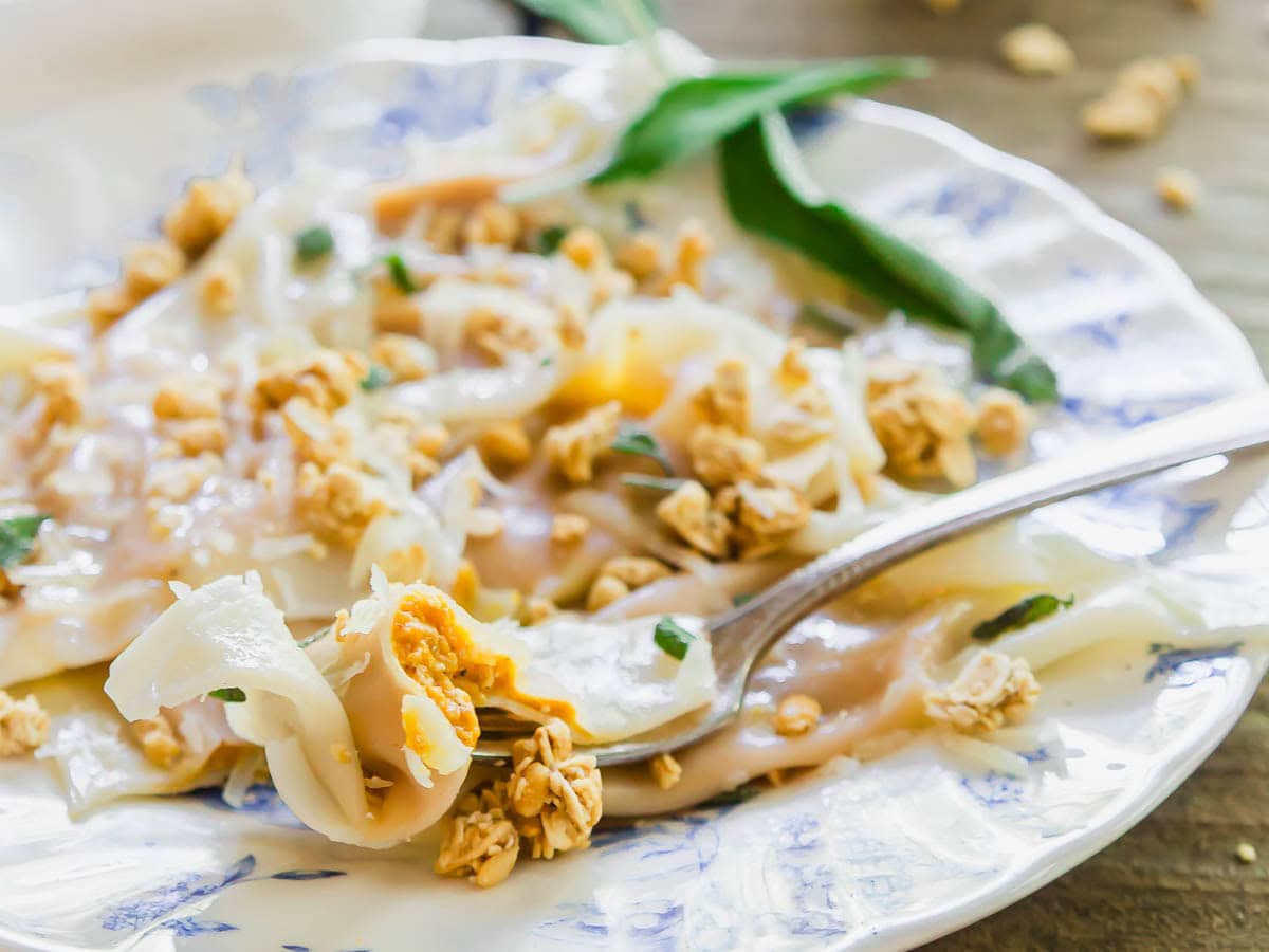 A close-up of a fork lifting a bite of thin rice noodle rolls topped with crushed peanuts and herbs on a patterned white plate.