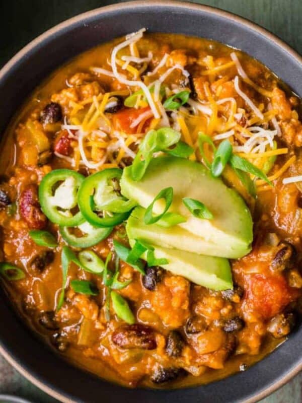 A bowl of chili topped with sliced avocado, shredded cheese, jalapeño, and green onions sits on a green table, surrounded by more bowls of chili, a small bowl of green onions, and a spoon.