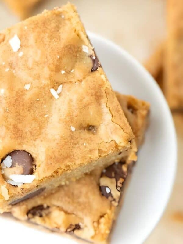Two blondie dessert bars with chocolate chips and sprinkled sea salt stacked on a white plate, with more blondie bars blurred in the background.