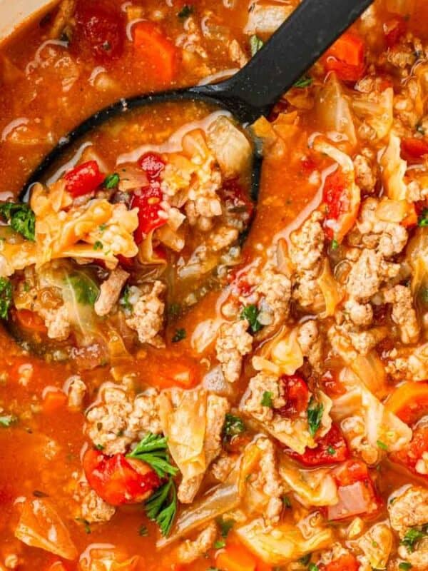 A pot of hearty soup with ground meat, chopped cabbage, tomatoes, onions, and herbs, with a black ladle lifting a serving. Slices of bread and a small bowl of pepper are nearby on a tiled surface.