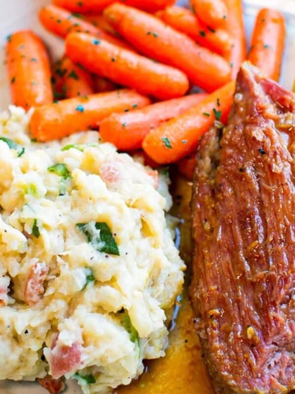 A plate with glazed baby carrots, two slices of tender pot roast with gravy, and a serving of chunky mashed potatoes with herbs and bacon bits.