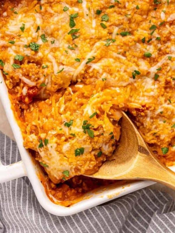 A baked spaghetti casserole topped with melted cheese and chopped parsley, with a wooden spoon lifting a serving from a white dish on a striped gray cloth.