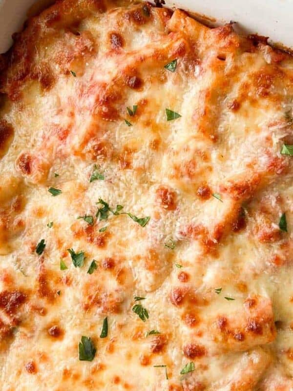 A baked pasta casserole topped with melted, golden-brown cheese and sprinkled with chopped parsley, in a white baking dish next to a wooden spoon and fresh parsley.