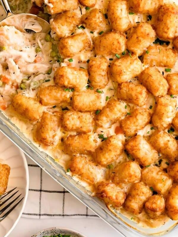A glass baking dish filled with a tater tot casserole, with a serving scooped onto a plate beside it. The casserole is topped with golden-brown tater tots and garnished with green herbs.