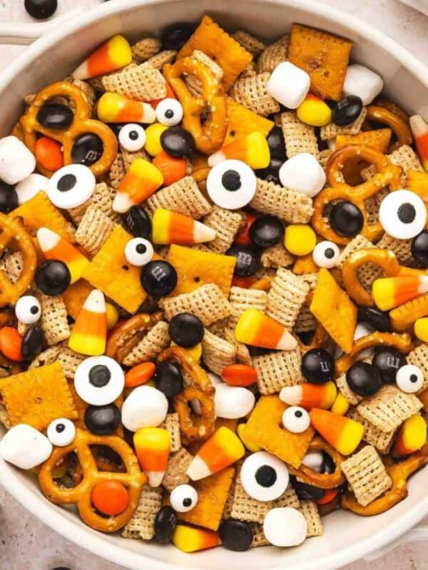 A bowl of Halloween snack mix with pretzels, Chex cereal, candy corn, chocolate candies, marshmallows, and candy eyeballs. Decorative toy ghosts, spiders, and marshmallows surround the bowl.