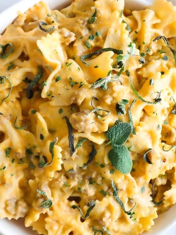 A bowl of farfalle pasta coated in a creamy orange sauce, garnished with chopped herbs and sage leaves, placed on a white surface next to a woven mat and some green plants.