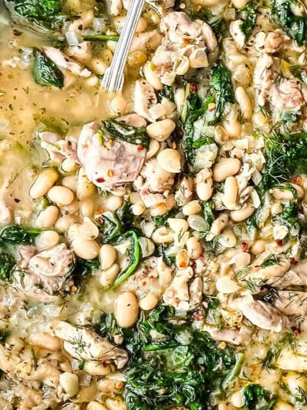 A skillet filled with a creamy stew of white beans, chicken pieces, wilted greens, and fresh herbs. A spoon rests in the dish, and a loaf of bread sits nearby on a wooden surface.