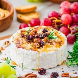 A wheel of honey baked brie topped with chopped pecans, dried cranberries and rosemary sits on parchment paper, surrounded by green and red grapes, crackers, and fresh rosemary.