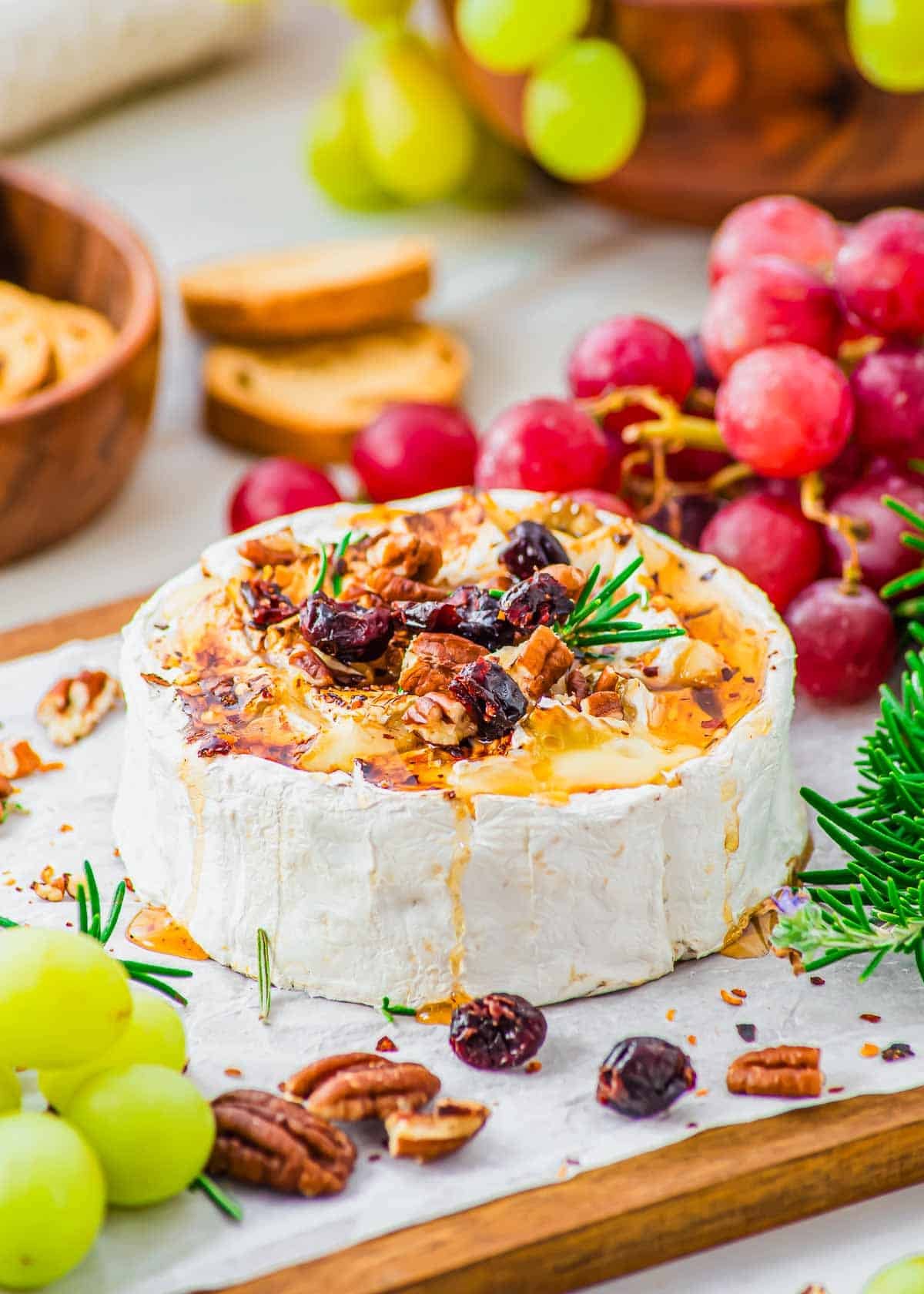 A wheel of honey baked brie topped with chopped pecans, dried cranberries and rosemary sits on parchment paper, surrounded by green and red grapes, crackers, and fresh rosemary.