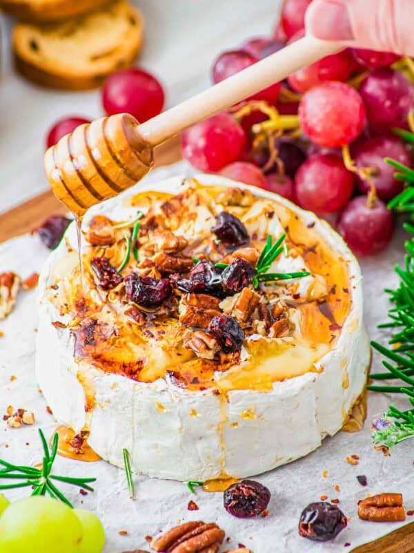 A wheel of baked brie topped with honey, chopped pecans, dried cranberries, and rosemary, being drizzled with more honey. Red grapes, green grapes, rosemary sprigs, and crackers surround the cheese on a parchment-lined board.